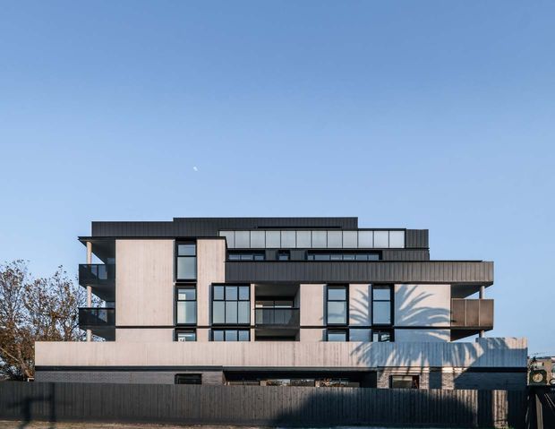 Exterior view of a contemporary multi-storey residential building with a strongly horizontal composition, dark ribbed cladding, pale vertical wall panels, extensive black-framed glazing, projecting balconies, and layered flat roof forms. The building is photographed from the street against a clear sky, with a front boundary fence and surrounding trees visible.