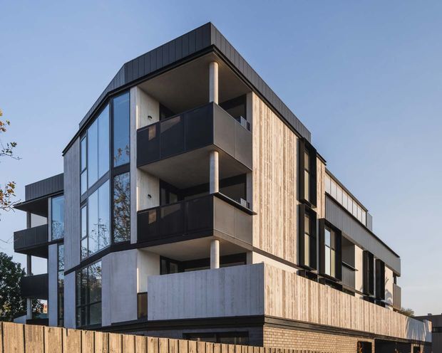 Exterior architectural photograph of a contemporary multi-storey apartment building with dark metal cladding, pale vertical timber-like wall cladding, concrete surfaces, expansive glazing, stacked balconies, and a flat roof.