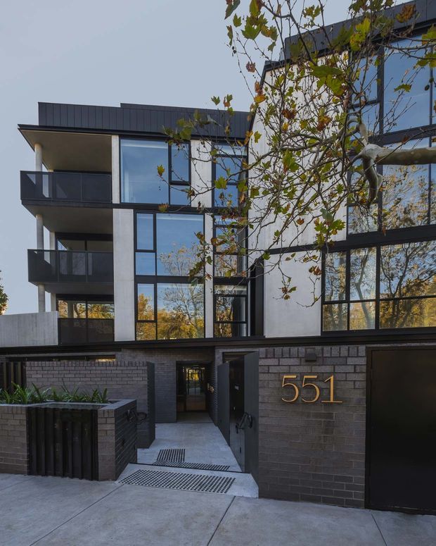Street-level exterior view of a contemporary multi-storey residential building. The facade combines dark brickwork, white rendered walls, extensive black-framed glazing, projecting balconies with dark balustrades, and a flat roofline. A gated pedestrian entry and large metallic street number are visible in the foreground, with mature trees reflected in the windows.