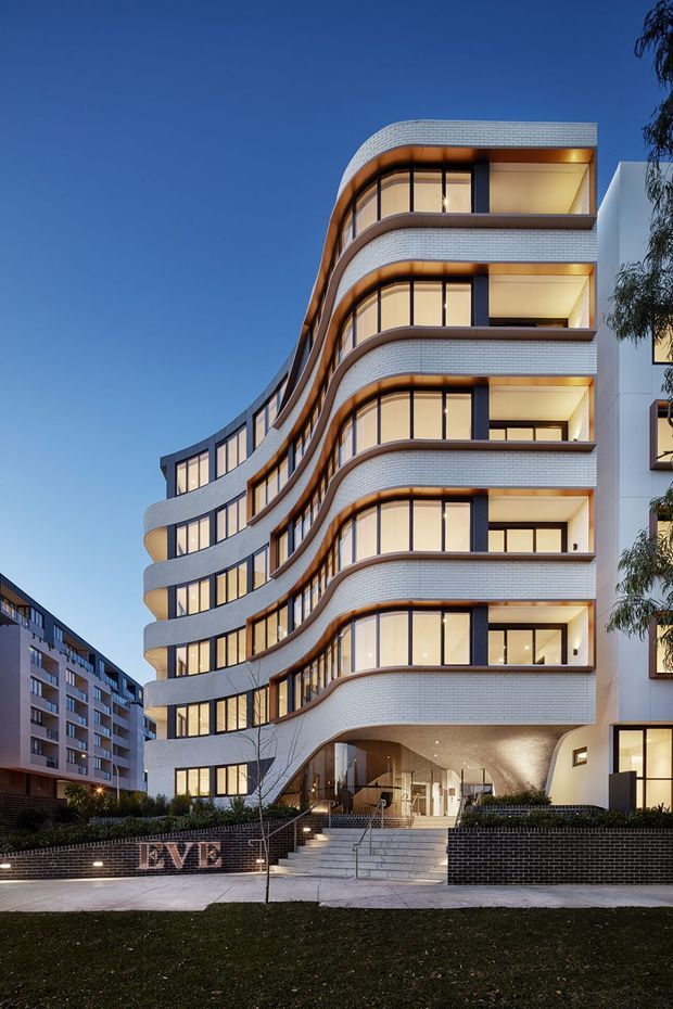 Twilight exterior view of a contemporary multi-storey apartment building with a sculptural, undulating facade. The building features curved projecting balconies, extensive floor-to-ceiling glazing, white textured brick or tile-like cladding, dark window frames, warm interior lighting, and a recessed entry beneath a cantilevered upper section. Landscaped frontage, steps, and neighbouring apartment buildings are visible.