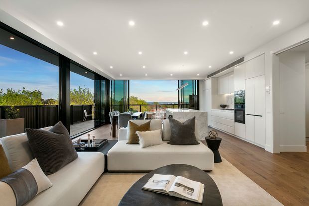 Open-plan contemporary living area with a large cream sectional sofa, dark round coffee table, and neutral rug, flowing into a compact kitchen with white floor-to-ceiling cabinetry and a pale stone island. Full-height black-framed glazing opens toward a balcony or terrace with greenery and distant views. Recessed ceiling lights, timber-toned flooring, and dark window frames create a refined, bright interior.