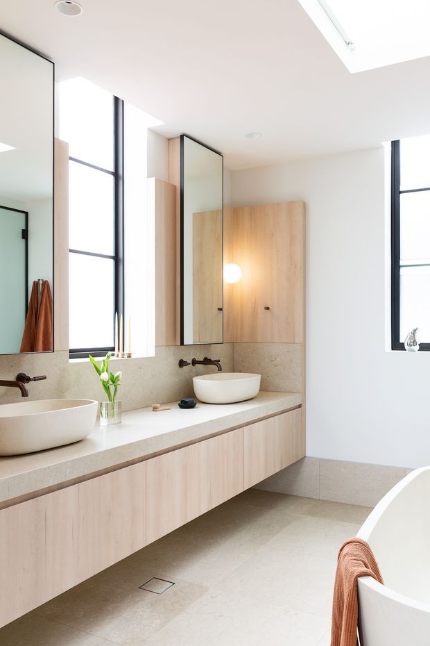 Bright, contemporary bathroom with a long floating timber vanity, pale stone benchtop, two white vessel basins, wall-mounted dark metal tapware, large mirrors, and tall black-framed windows. A freestanding white bathtub is partially visible in the foreground. The room features pale natural finishes, integrated cabinetry, skylight-like ceiling glazing, and warm accent lighting.