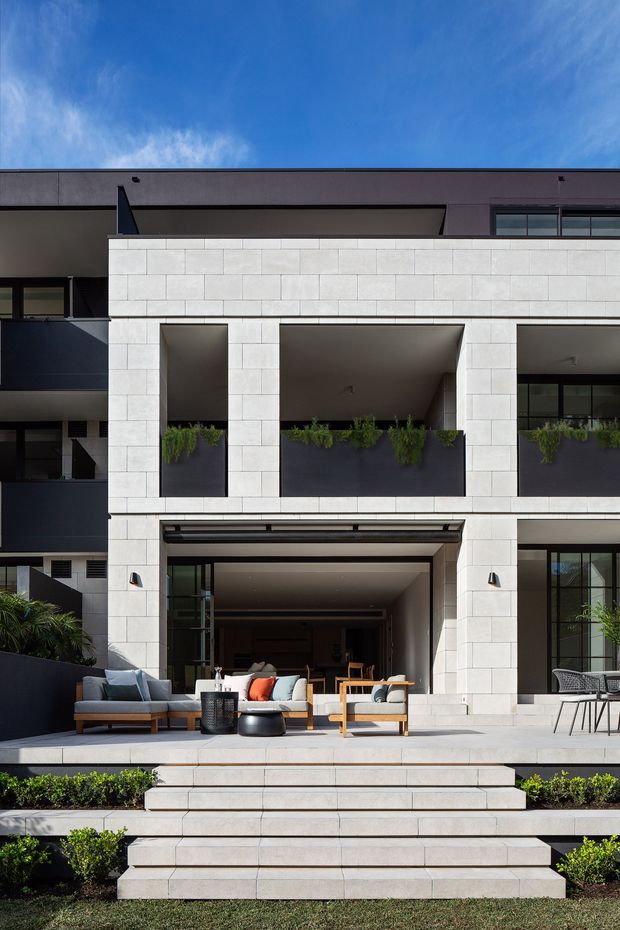 A contemporary multi-storey residential facade with a broad outdoor terrace and recessed covered entertaining area. The building features a rectilinear composition, dark upper-level walls and window frames, pale large-format rectangular masonry or stone cladding, deep square openings, integrated planter boxes with trailing greenery, and wide tiled steps leading down to a landscaped lawn. Outdoor lounge furniture is arranged beneath the covered terrace.