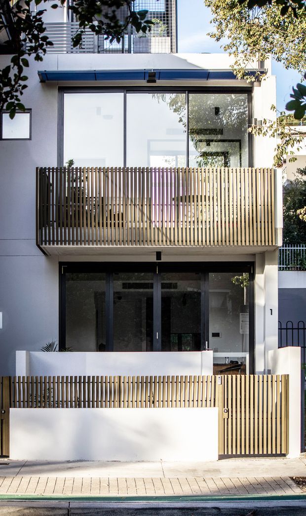 Front exterior view of a narrow multi-storey contemporary townhouse or apartment-style residence. The white rendered facade has large dark-framed glazed openings, projecting blue-grey metal awnings, and two levels of private balconies screened by closely spaced vertical timber battens. A dark glazed entry is visible at ground level, with a small paved frontage and street edge in the foreground. Mature foliage partially frames the building.