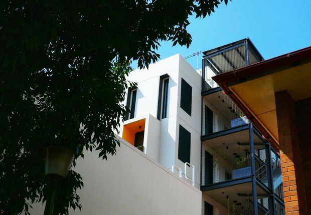 Exterior architectural view of a contemporary multi-storey building, partially framed by a leafy tree. The white rendered facade has sharp geometric forms, tall dark window openings, and an orange recessed entry or balcony. To the right is a metal-framed, mesh-enclosed external stair or balcony structure, adjoining a brick building with a projecting red-edged roof.