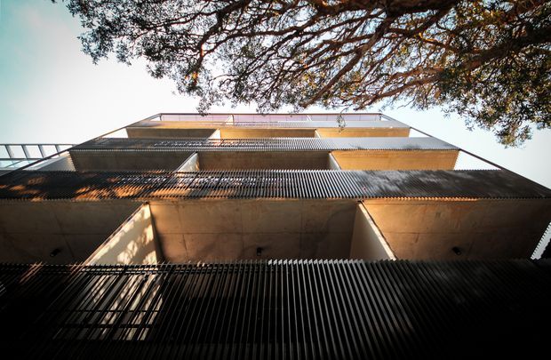 Upward-looking architectural photograph of a multi-storey contemporary building facade framed by overhanging tree branches. The facade features layered projecting balconies or terraces, extensive dark vertical metal screening, pale rendered or concrete wall planes, and horizontal upper-level glazing and railings. Strong sunlight creates patterned shadows across the building.