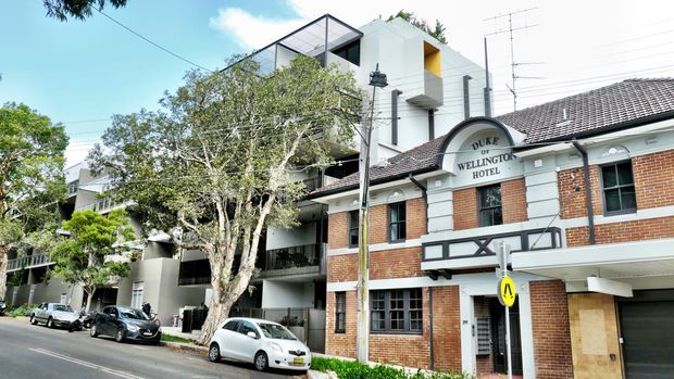 Street-level exterior view of the Duke of Wellington Hotel, a heritage-style red-brick building with white-painted trim, arched signage, tiled roof and projecting balconies. A contemporary multi-storey apartment or commercial building is visible immediately behind it, with mature street trees, parked cars and urban streetscape elements.