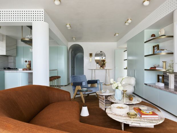 A refined open-plan living room with a warm brown curved sofa in the foreground, layered round coffee tables, sculptural lounge chairs, and built-in pale blue-grey shelving and cabinetry. The space connects to a compact kitchen and dining/bar area, with arched openings, fluted wall detailing, perforated decorative ceiling bands, dark timber flooring, marble-patterned table surfaces, and polished brass accents. The overall interior is bright, sophisticated, and contemporary with Art Deco-inspired detailing.