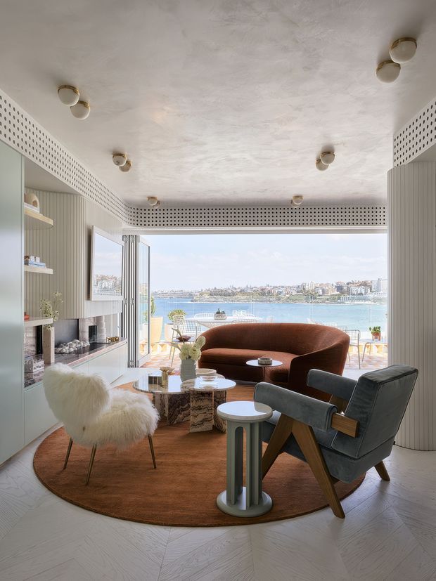 A bright, curated living room overlooking a blue waterfront through wide open glazing. The room features a sculptural brown curved sofa, dark grey lounge chair, fluffy white accent chair, round coffee tables, a large rust-toned circular rug, pale herringbone timber flooring, fluted wall panelling, open shelving, and a textured white ceiling with perforated trim detailing. A built-in fireplace and television wall occupy the left side, while the outdoor terrace and waterfront are visible beyond.