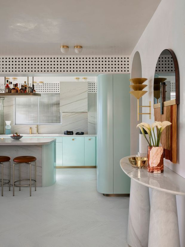 A refined retro-inspired open-plan kitchen and bar area with pale mint cabinetry, a curved island, integrated appliances, and a perforated decorative wall band. The space features a white and grey veined stone-look palette, rounded architectural forms, brass accents, sculptural lighting, arched mirrors, and a curved console displaying flowers and decorative objects.