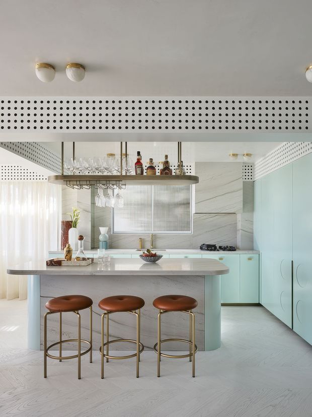A bright, refined kitchen with pale aqua cabinetry, a large marble island, and three brown leather bar stools. A suspended circular drinks shelf with hanging glassware forms a feature above the island. The space includes a marble-look splashback and wall finish, pale timber flooring, perforated white architectural detailing across the ceiling bulkhead and upper walls, and brass-toned lighting and hardware.