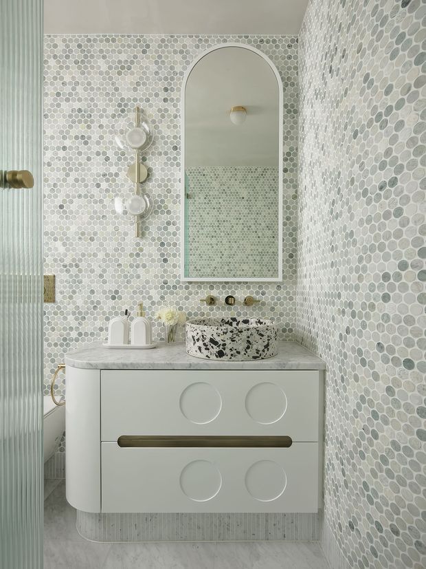 A compact, refined bathroom vanity viewed straight on. The room is wrapped in small pale grey and white mosaic tiles, with a rounded-arch mirror, wall-mounted circular lights, a white floating vanity with sculptural circular drawer details, a long brass-toned handle, a pale stone benchtop, and a terrazzo vessel basin. A fluted translucent glass door or partition is visible at left.