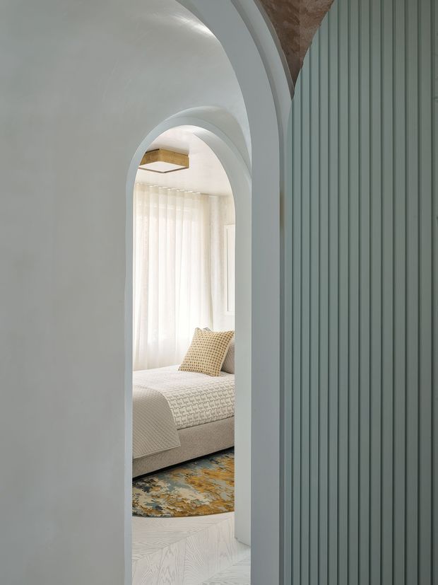 A softly lit architectural interior view through a tall rounded archway into a serene bedroom. The foreground features a muted blue-grey vertical fluted wall or fixed paneling, while the bedroom beyond has a light upholstered bed, textured neutral bedding, sheer curtains, a patterned blue-and-gold rug, and warm timber accents. Curved plasterwork and arched openings create a refined, sculptural transition between spaces.