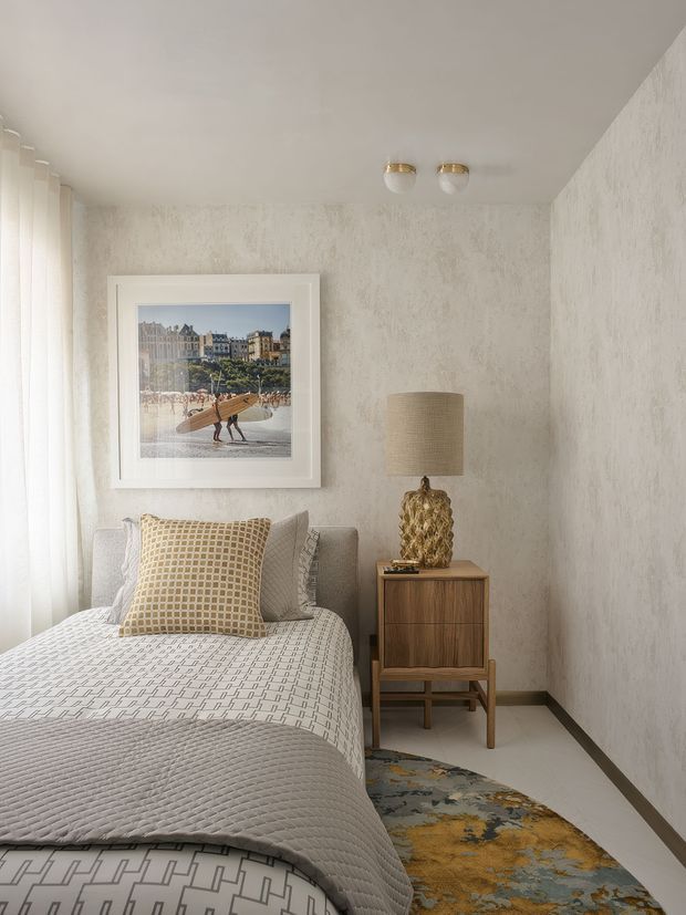 A compact, softly lit bedroom with a single bed dressed in layered white and grey geometric bedding, textured off-white walls, sheer curtains, and a warm timber bedside cabinet. A sculptural lamp with a woven shade sits beside the bed, while framed coastal artwork and a patterned rug add subtle colour and texture. The overall look is refined, calm, and contemporary with tactile finishes.
