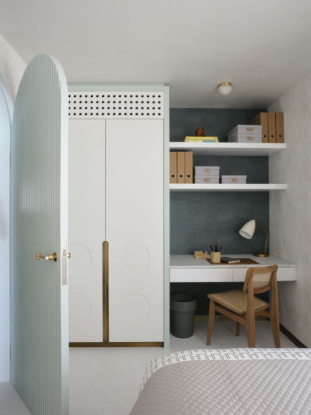 Compact, refined bedroom nook with a built-in home office and wardrobe. The space features a pale ribbed arched door, white custom cabinetry with circular detailing and brass handles, open shelving with organized storage boxes, a dark blue-grey textured wall behind the desk, a woven wooden chair, desk lamp, and light patterned flooring. The foreground shows part of a neatly made bed.