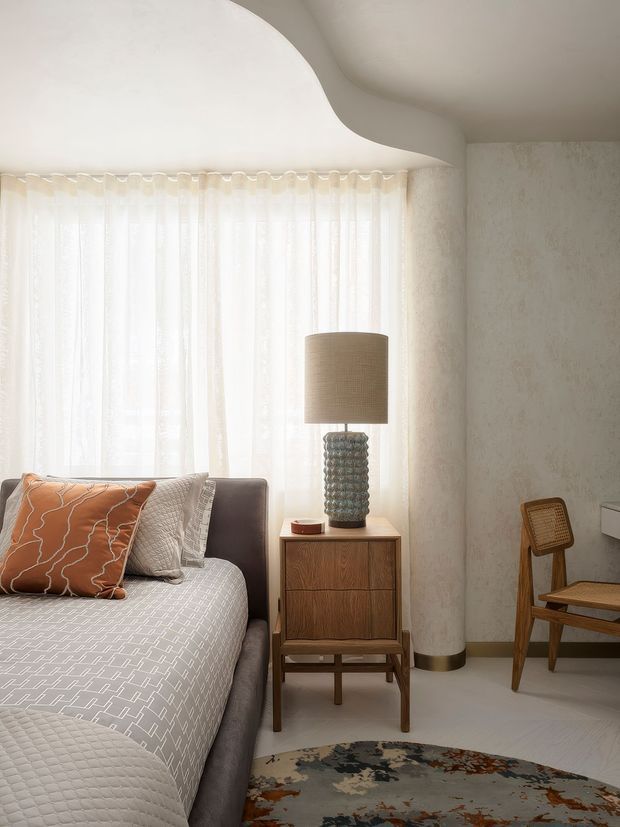 A softly lit, refined bedroom interior with a curved sculptural ceiling, sheer full-height curtains, textured plaster-like walls, and pale flooring. A bed with a geometric patterned cover and warm orange accent cushion sits beside a mid-century timber bedside cabinet, ceramic-textured lamp, cane chair, and muted floral rug. The palette is warm neutral with restrained earthy accents.
