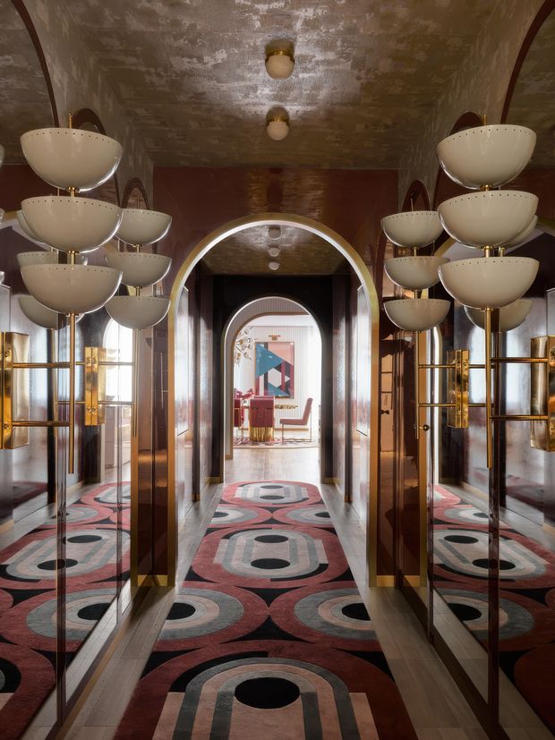A dramatic, symmetrical Art Deco-style interior hallway or entry corridor with mirrored walls, richly patterned geometric carpet, dark wood or lacquered wall panels, and multiple sculptural tiered wall sconces in cream and brass. Repeated arched openings lead toward a bright sitting area, creating a strong sense of depth and reflection.