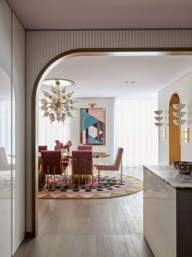 A refined dining room viewed through a large rounded archway, featuring vertically ribbed wall panelling, sheer floor-to-ceiling curtains, a sculptural brass-and-glass chandelier, burgundy upholstered dining chairs, a geometric multicoloured rug, and a round dining table. The foreground includes a marble-topped kitchen or bar counter, while brass accents and a bold abstract artwork add Art Deco character.
