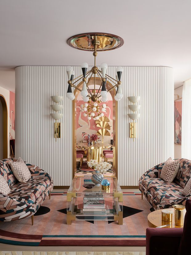 A richly styled, symmetrical living room with two patterned sofas facing a glass coffee table and central mirror. The room features a fluted white feature wall, ornate brass-and-glass chandelier, matching wall sconces, geometric patterned upholstery, colourful geometric artwork, and a striped geometric rug. The overall aesthetic is highly decorative, glamorous, and Art Deco-inspired, with warm brass accents and bold blush, teal, cream, and brown tones.