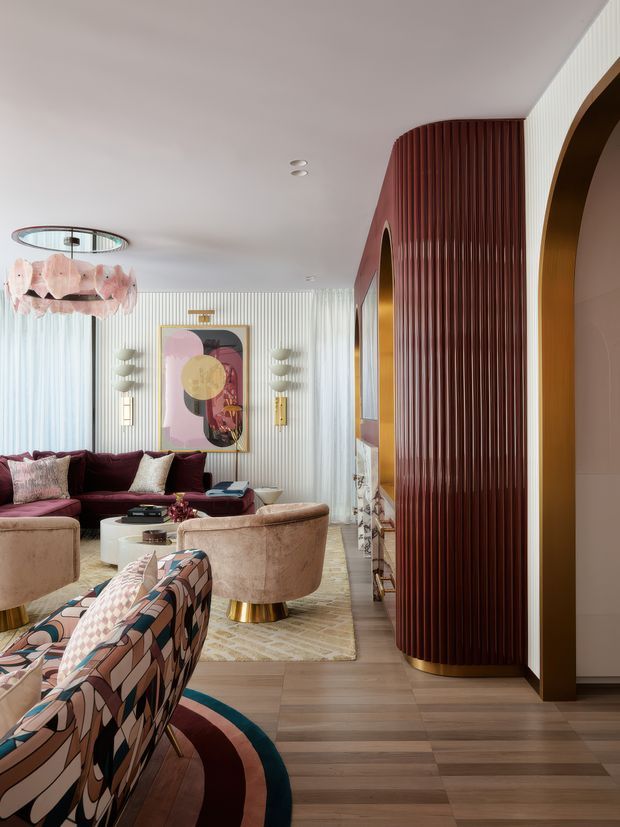 A richly styled living room with a curved burgundy sofa, plush blush-toned armchairs, patterned upholstered seating, and a round coffee table. The room features warm timber flooring, a large cream rug, vertical fluted wall panelling, a dramatic curved burgundy ribbed feature wall with brass-toned base detailing, arched openings, sculptural wall sconces, and a decorative pendant light. The palette is warm, layered, and glamorous with strong geometric and organic forms.