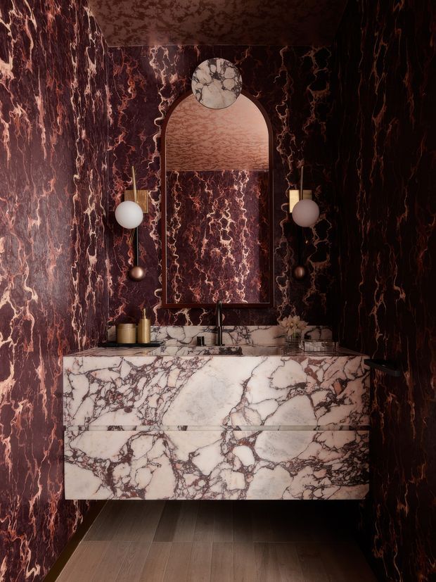 A dramatic, compact bathroom vanity vignette clad almost entirely in richly veined burgundy and plum marble. A floating double-drawer marble vanity supports a pale stone benchtop with an integrated basin and dark metal tapware. Above it is an arched mirror with a circular decorative marble detail, flanked by globe wall lights. The dark timber-look flooring and deep tones create a moody, luxurious atmosphere.