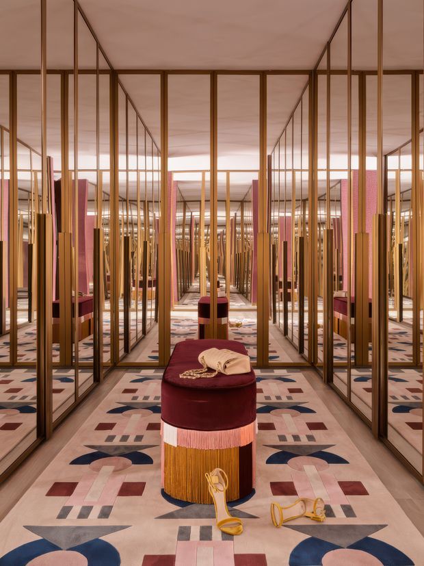 A highly stylised, symmetrical interior featuring mirrored floor-to-ceiling wall panels framed in warm bronze or brass-toned metal. Repeated reflections create a long, immersive corridor-like space. The room has a pale pink ceiling and a boldly geometric patterned carpet in blush, burgundy, navy, cream, and taupe. A central cylindrical burgundy upholstered ottoman with fringed and metallic detailing displays folded textiles and accessories, giving the setting a luxurious boutique or showroom character.