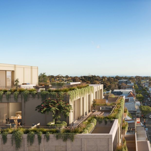 A contemporary multi-storey residential building viewed from an elevated exterior vantage point, featuring extensive planted terraces, rooftop gardens, concrete-clad walls, vertical screening, balconies and large glazed openings. A suburban neighbourhood and distant coastal horizon are visible beyond the building.