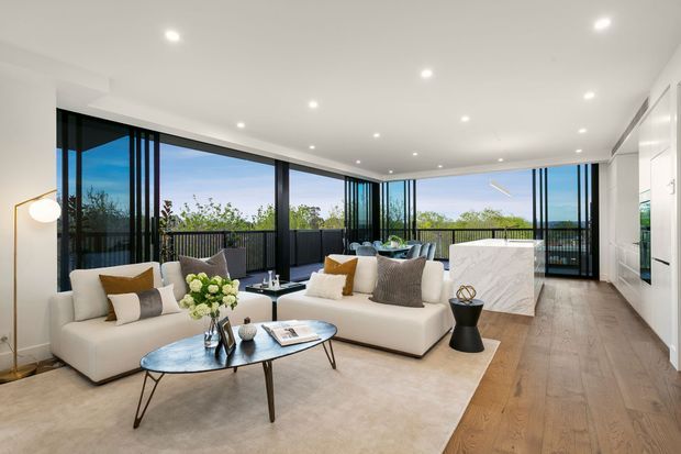 Bright, spacious contemporary living room with floor-to-ceiling black-framed glazing opening onto a balcony or terrace. The room features white sofas, a marble-clad rectangular feature island or bar, a large neutral rug, timber flooring, recessed ceiling lights, and minimal modern furnishings. An integrated kitchen or joinery wall is visible at the right.