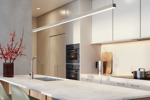 A refined contemporary kitchen featuring a large pale stone island with integrated double sink and tall curved faucet, full-height light cabinetry, stacked black appliances, a pale splashback, and a linear suspended pendant light. A dark vase with red blossom branches provides a small decorative accent.
