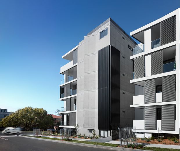 Exterior view of a contemporary multi-storey apartment building with a light concrete central facade, dark vertical screening, projecting balconies, white horizontal slab edges, and louvred window shutters. A second adjoining modern apartment wing is visible on the right, with landscaped planting, pedestrian paths, and a residential street in the foreground.