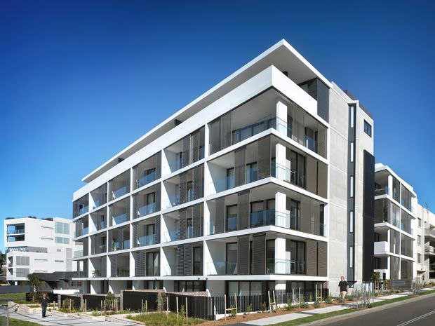 Exterior architectural photograph of a contemporary multi-storey apartment building with crisp white horizontal slab edges, extensive floor-to-ceiling glazing, recessed balconies, dark vertical screening elements, and a flat roof. The building fronts a paved street with landscaped pedestrian edges under a clear blue sky.