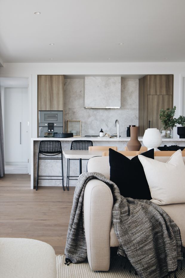 A bright open-plan kitchen and living area with a neutral contemporary fit-out. The kitchen features tall timber-grain cabinetry, a pale stone island and splashback, integrated appliances, and black metal bar stools. In the foreground is a light upholstered sofa with black and cream cushions and a textured grey throw, set over light timber flooring. Recessed ceiling lights and restrained styling create a clean, refined atmosphere.