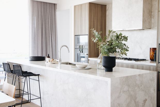 Bright contemporary kitchen with a large pale stone island and waterfall edge, integrated sink and curved tap, black bar stools, timber-toned tall cabinetry, light marble-effect splashback, and a sculptural white rangehood. Soft grey curtains and natural greenery add warmth to the refined, neutral space.
