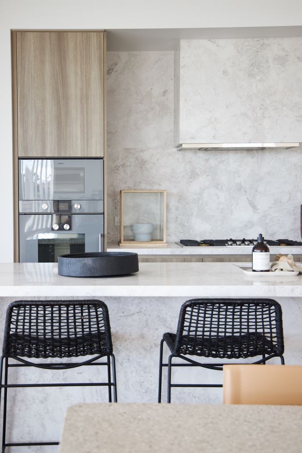 A refined contemporary kitchen viewed from the dining area, featuring a large pale stone island with waterfall edge, integrated light timber cabinetry, built-in stainless-steel ovens, a pale veined stone splashback and rangehood surround, and a gas cooktop. Two black woven bar stools sit at the island, with a dining table in the foreground. The palette is soft grey, white, light timber and black, creating a clean, understated look.
