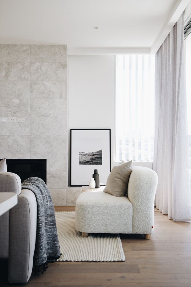 Bright, refined living room corner with a sculptural cream boucle lounge chair, neutral cushion, textured rug, pale timber flooring, full-height sheer curtains, and a grey-toned stone or marble fireplace wall. A framed monochrome artwork and small decorative vessels sit beside the fireplace, while part of a grey sofa is visible in the foreground.