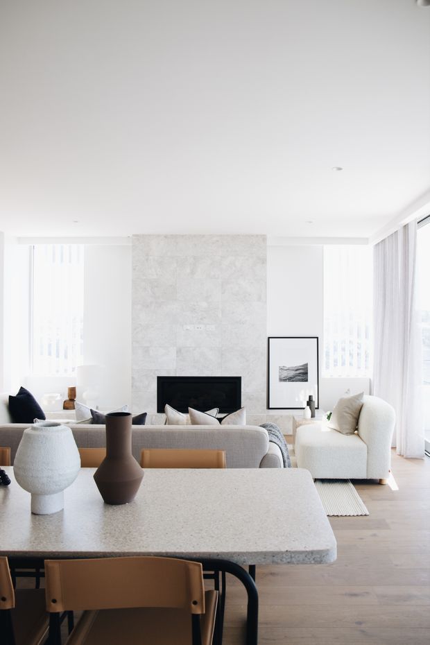 Bright, refined open-plan living and dining space with a large pale stone dining island or table in the foreground, leather dining chairs, a neutral sofa, sculptural armchair, and a black-framed fireplace set into a light textured stone feature wall. Full-height sheer curtains and glazing bring in abundant daylight, while pale timber flooring and restrained decor create a calm contemporary interior.