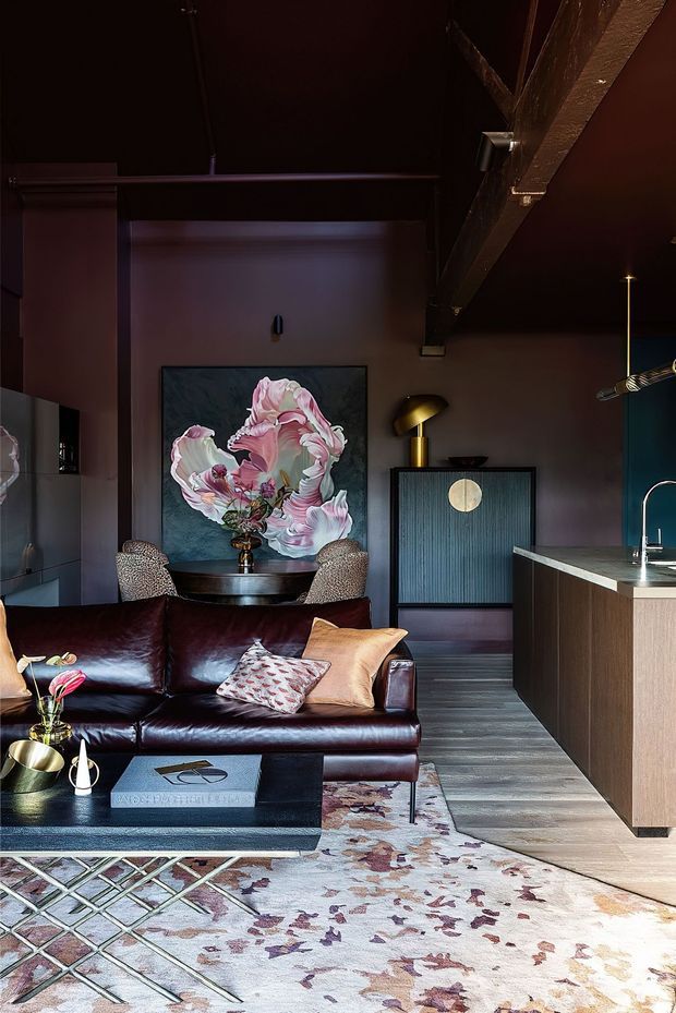 A richly styled open-plan living room with a dark leather sectional, patterned rugs, sculptural coffee table, floral artwork, and a compact dining setting. A contemporary kitchen with a wood-front island and integrated sink is visible to the right. The room has a dark, atmospheric palette, exposed structural beam, layered designer lighting, and a dramatic floral feature artwork.