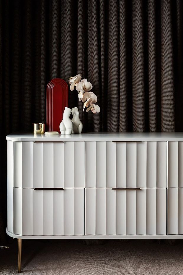 A refined interior vignette featuring a curved white sideboard with vertically fluted drawer and door fronts, slim dark metal legs, and a pale smooth countertop. A dark textured, vertically pleated wall covering forms the backdrop, styled with white ceramic vases, a red arched decorative panel, an orchid arrangement, and small metallic accessories. The composition has a sophisticated, high-contrast contemporary look.