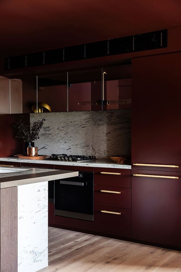 A dark, dramatic contemporary kitchen with deep burgundy cabinetry, slim brass handles, a pale stone benchtop and matching textured stone splashback. A black cooktop and oven sit beneath mirrored upper cabinetry, while a large island with a pale stone waterfall end and timber side panel occupies the foreground. Timber flooring and warm low lighting create a refined, moody atmosphere.