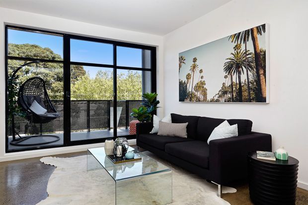 Bright contemporary living room with a black sofa, glass coffee table, sculptural black side table, and a large palm-tree artwork. Floor-to-ceiling black-framed glazing opens to a balcony with greenery beyond, while a hanging egg chair sits by the window. The room features white walls, a polished concrete floor, and a light marble-look rug.