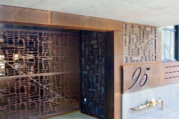 An architectural exterior entry or lift lobby featuring weathered steel framing and decorative perforated metal screens with organic, rounded-slot patterns. A dark glazed doorway or elevator entrance is set within the rust-toned metal surround, with a stylised number or letter graphic on the adjacent panel. Exposed concrete surfaces and service taps add an industrial character.