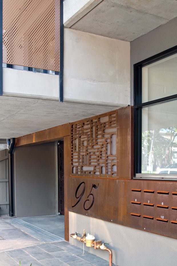 A close-up view of a modern building entrance or frontage beneath a concrete canopy. Rust-toned perforated metal screens and cladding form a decorative security and mailbox panel, marked with the number 93. Black-framed glazing reveals a bright interior, while exposed plumbing and dark paving contribute to an industrial architectural character.