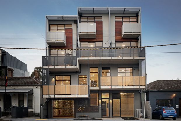 Street-facing view of a contemporary multi-storey apartment building with a strong rectilinear facade, dark metal balcony and terrace balustrades, large glazed openings, projecting balconies, and vertical timber-toned screening panels. The building is closely flanked by older low-rise homes in an urban residential streetscape.