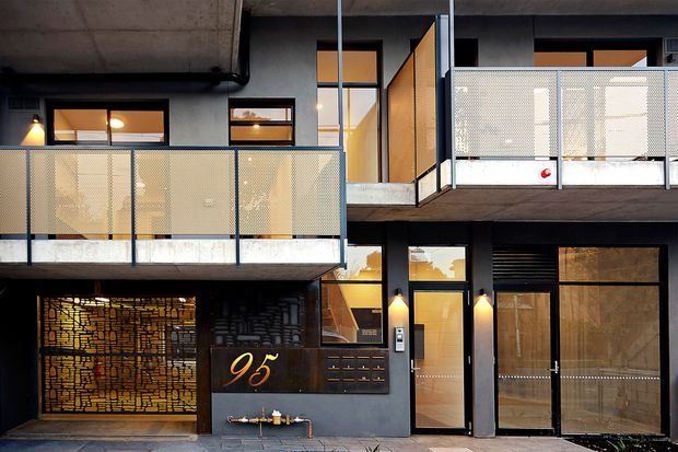 Exterior view of a contemporary multi-storey residential building with dark concrete and masonry surfaces, warm-lit windows, projecting balconies, and perforated metal privacy screens. The ground level includes a recessed entry or garage-like opening with decorative patterned screening, a dark framed entrance door, wall-mounted lighting, and the illuminated house number 95.