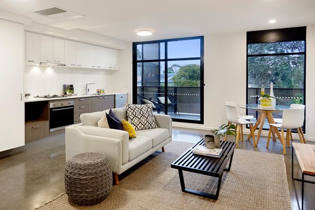 Open-plan contemporary apartment interior combining a compact white and grey kitchen with a lounge and dining area. The space features full-height black-framed glazing, polished concrete-look flooring, a neutral woven rug, a cream sofa, black slatted coffee table, and a light timber dining setting. The large windows provide abundant natural light and views to greenery and neighboring buildings.