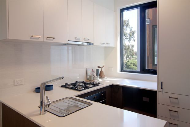 Compact contemporary kitchen with an L-shaped white benchtop, integrated sink and chrome tap, gas cooktop, dark lower cabinetry, white upper cabinetry, and a glossy light tiled splashback. A large black-framed window provides natural light and views of greenery.