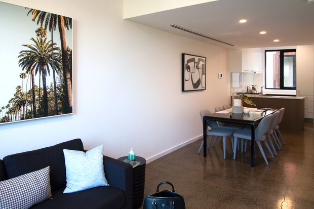 Open-plan contemporary apartment interior combining a compact living area, dining zone, and kitchen. A dark sofa with patterned cushions sits beside a large palm-tree artwork, while a dark dining table with upholstered chairs leads toward streamlined cabinetry, stainless appliances, and a window. Polished concrete flooring, recessed lighting, and a neutral palette create a clean, modern presentation.