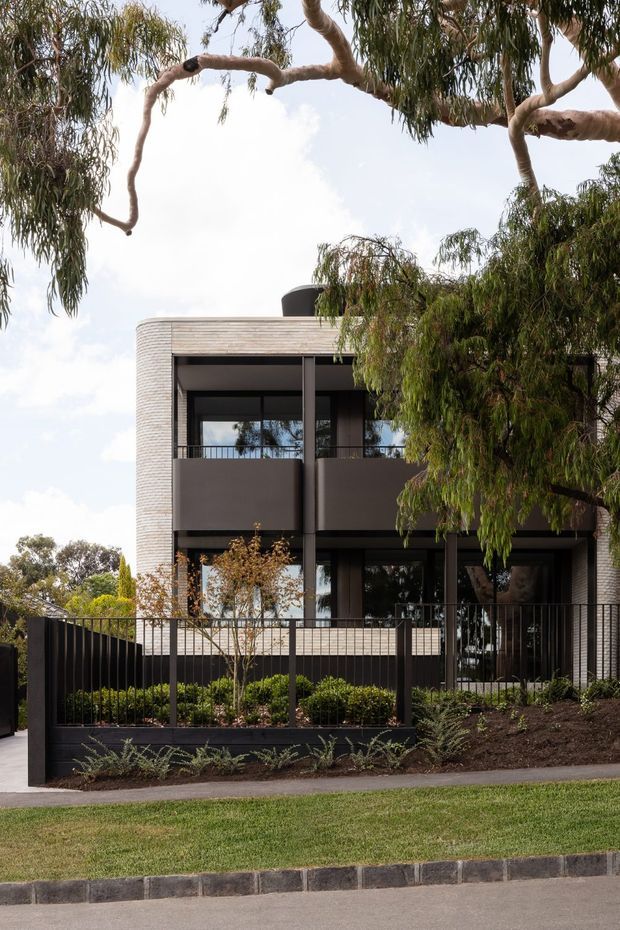 Two-storey contemporary residential facade viewed from the street, framed by mature eucalyptus trees. The house has rounded corners clad in light horizontal brickwork, dark metal-framed glazing, recessed balconies, dark privacy screens and a black metal front fence. A landscaped front garden, lawn and paved street edge are visible.
