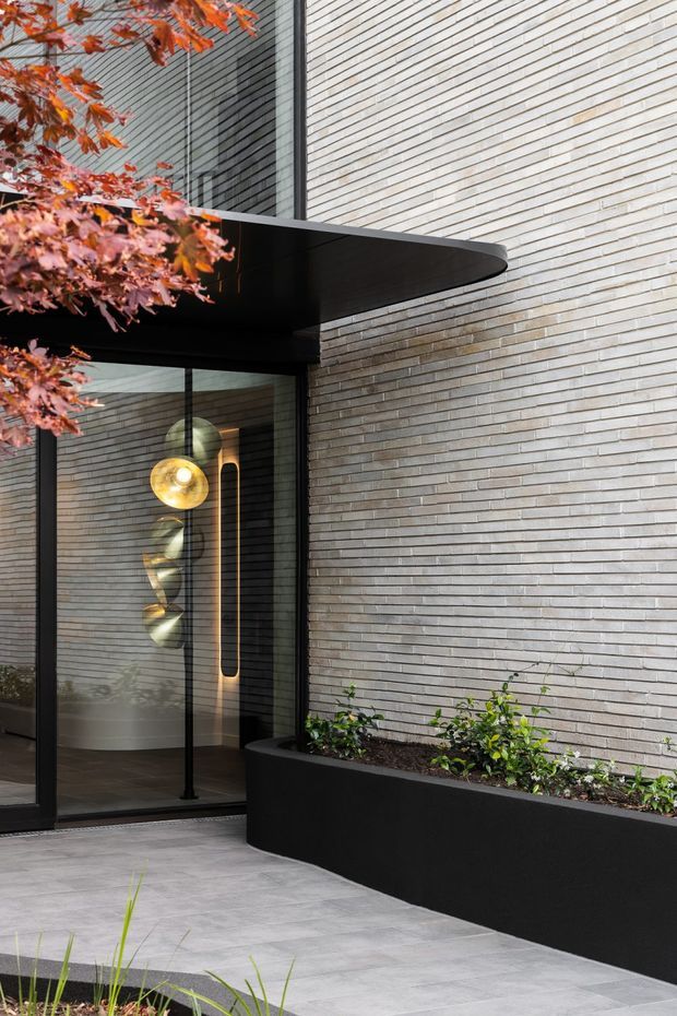 Contemporary residential entrance with a pale grey linear brick facade, dark glazed entry door, and a broad curved black canopy. A black built-in planter runs along the wall, while a sculptural pendant light is visible through the glazing. Grey tiled paving and clipped planting complete the refined exterior composition.