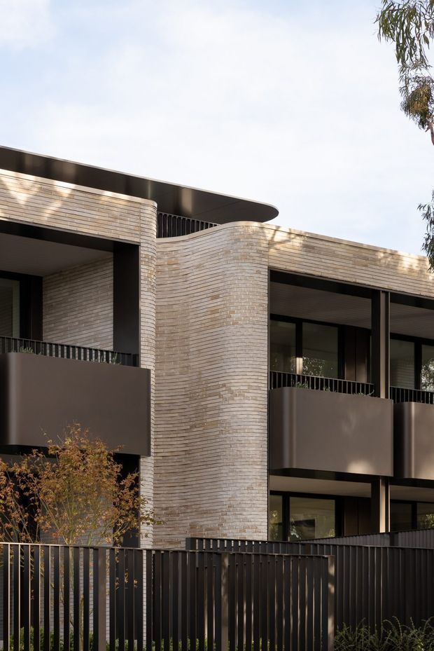 Exterior architectural view of a contemporary multi-storey residential building. The facade combines pale, narrow-format brickwork with rounded projecting wall forms, dark metal balcony balustrades and screening, large dark-framed glazing, and deep charcoal roof and balcony elements. A tall dark metal fence and sparse landscaping occupy the foreground. The building has a strong horizontal composition with curved corners and an overhanging, gently curved roof edge.